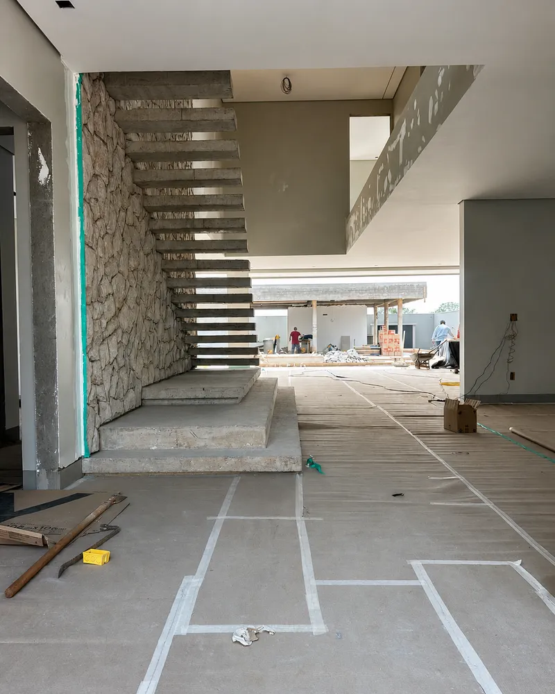 Interior de obra em fase de acabamento, com escada de concreto aparente e equipe trabalhando ao fundo